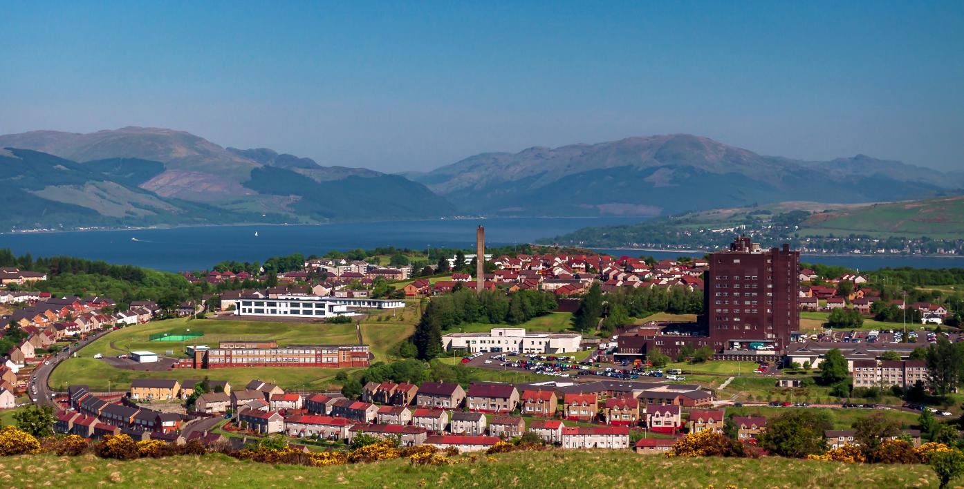 An aerial image of Inverclyde, featuring estates of red rooved houses on a green hill with the Firth of Clyde  in the back ground. 