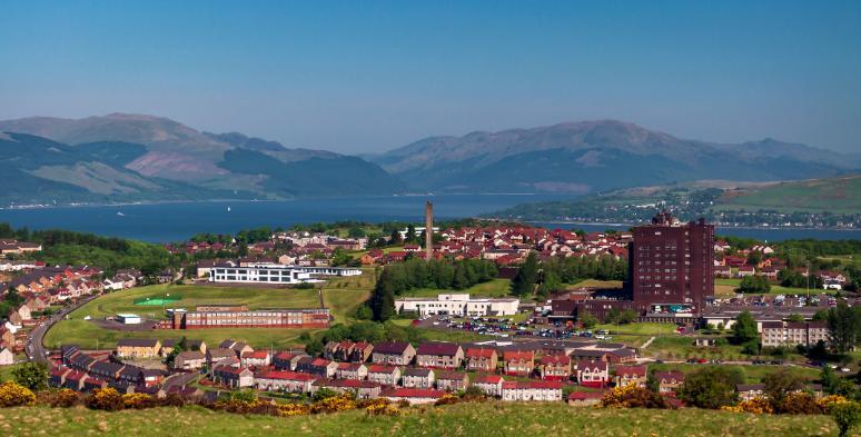 An aerial image of Inverclyde, featuring estates of red rooved houses on a green hill with the Firth of Clyde  in the back ground. 