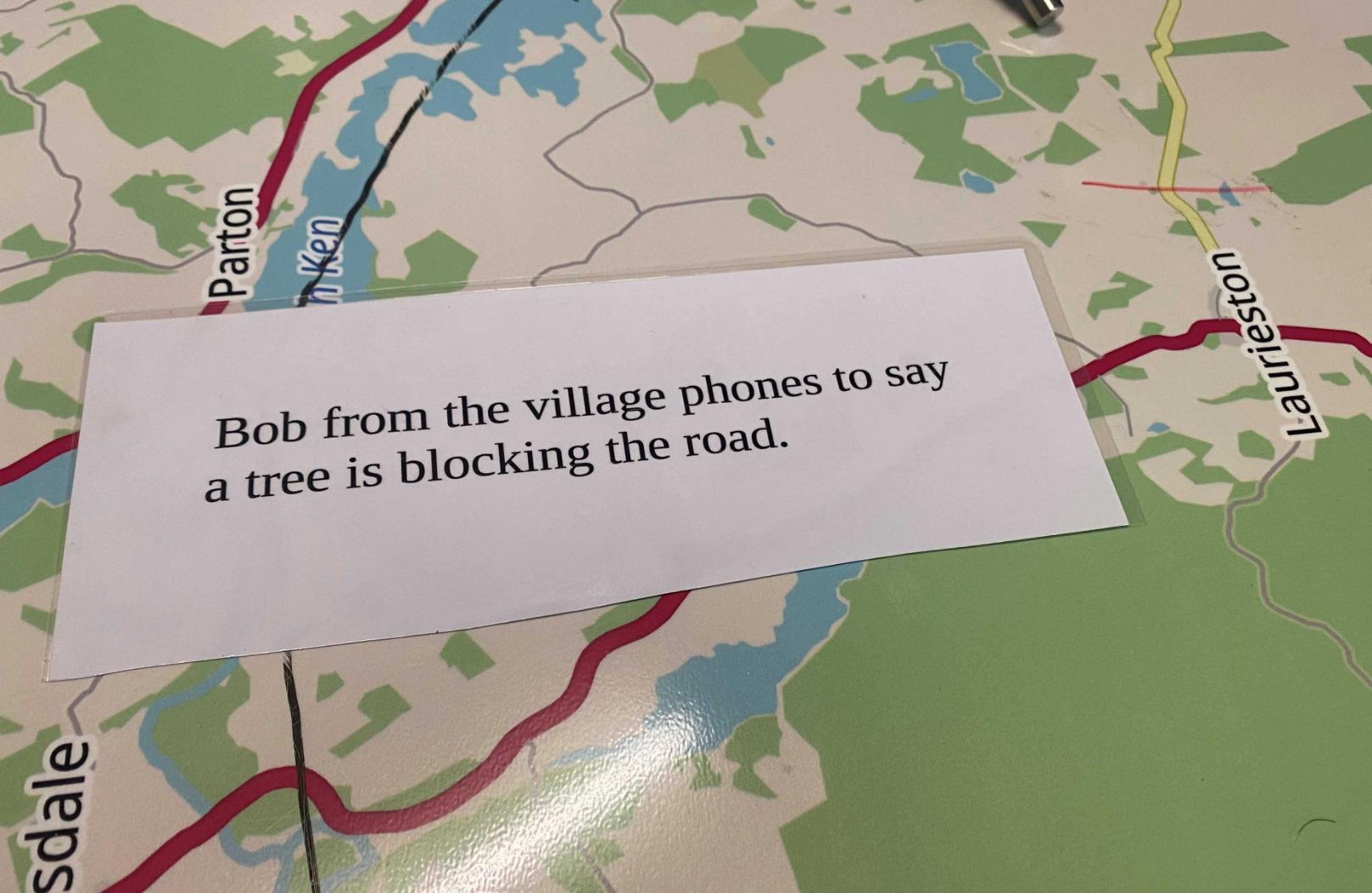 Image shows a scenario card laid over a map. The scenario says: Bob from the village phones to say a tree is blocking the road.