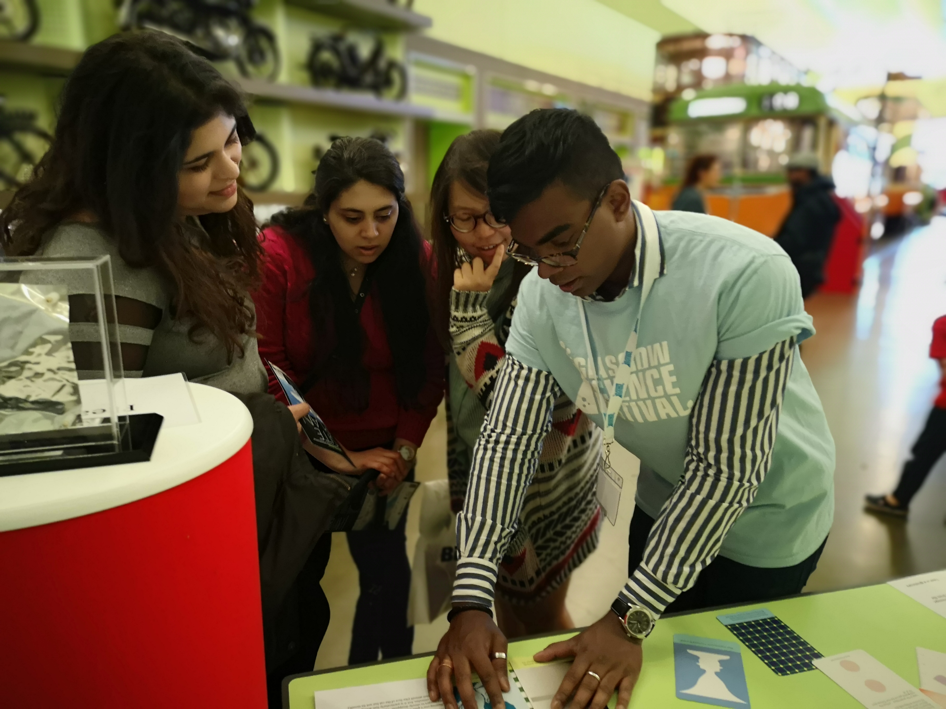 photograph of a phd student (a young black man with glasses and short hair) in a light blue t-shirt demonstrates examples of visual illusions to visitors at Glasgow Science Festival